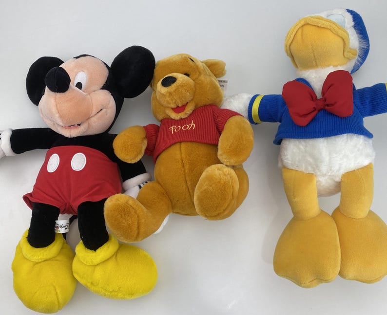 May include: Three plush Disney character toys: Mickey Mouse in red shorts, Winnie the Pooh in a red shirt, and Donald Duck in a blue sailor suit with a red bow tie. The toys are soft and colorful.