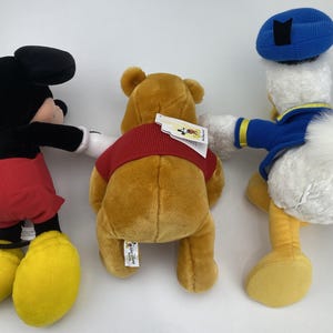 May include: Three plush Disney character toys: Mickey Mouse, Winnie the Pooh, and Donald Duck. Mickey Mouse is in a classic outfit, Winnie the Pooh wears a red shirt, and Donald Duck has a blue sailor hat and jacket.