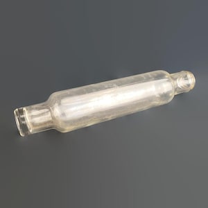 May include: A clear glass bottle, cylindrical in shape, with a narrow neck and a wider base. The bottle is empty and has a vintage look. It is lying horizontally on a dark grey surface. The bottle's length is approximately 20 cm.