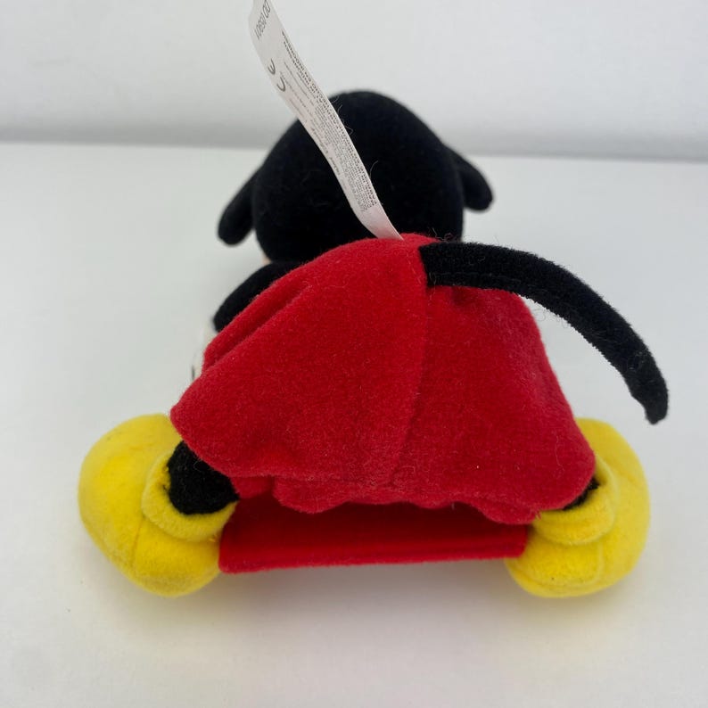 Puede incluir: Un peluche de un rat&oacute;n de dibujos animados, visto desde atr&aacute;s. El rat&oacute;n tiene orejas negras y una cola negra. Lleva pantalones cortos rojos y zapatos amarillos. Una etiqueta blanca est&aacute; adherida al juguete.