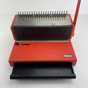 May include: A red Ibico binding machine featuring a metal punching plate with vertical teeth, a black pull-out tray at the front, and a red manual lever on the right side. The device includes a printed scale and the brand name Ibico on the front.
