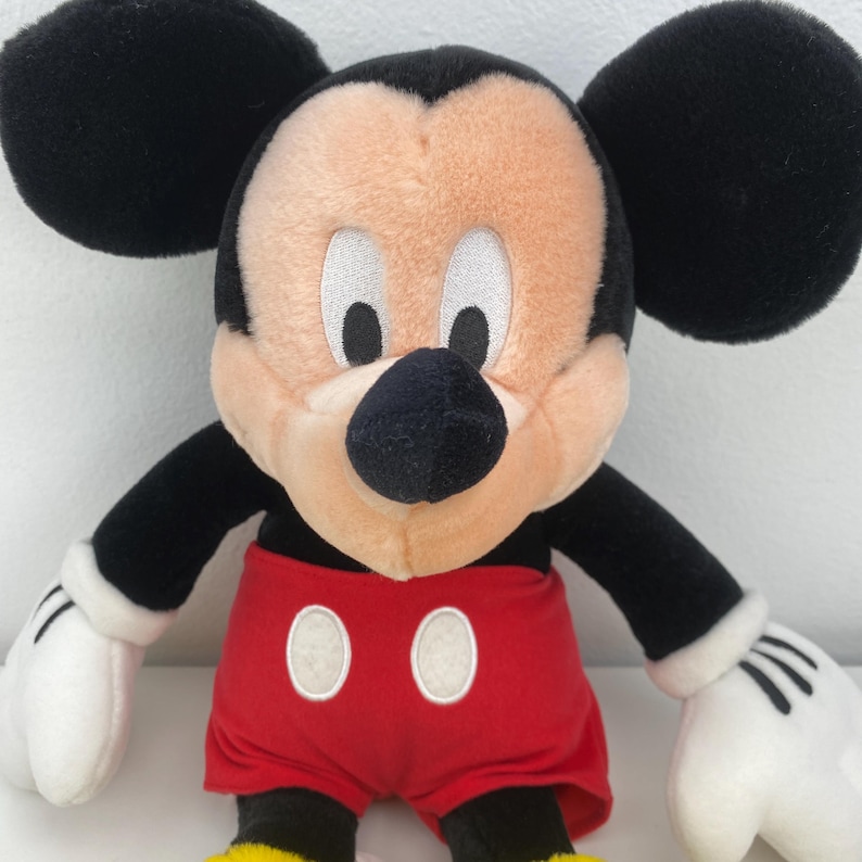 May include: A plush Mickey Mouse toy with black ears, a black nose, and a peach-colored face. The toy has black arms, white gloves, and red shorts with two white buttons. The toy is a classic Disney character.