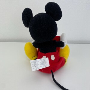Puede incluir: Un peluche de Mickey Mouse, visto desde atr&aacute;s, con orejas y cabeza negras, guantes blancos, pantalones cortos rojos con botones blancos y zapatos amarillos. Una cola negra se extiende desde los pantalones cortos. Una etiqueta blanca est&aacute; adjunta.