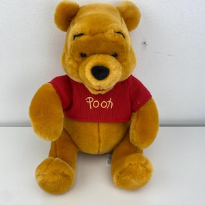May include: A plush Winnie the Pooh bear, featuring a golden-yellow fur and a red sweater. The sweater has the word "Pooh" embroidered in a gold color. The bear has a black nose and is sitting upright.
