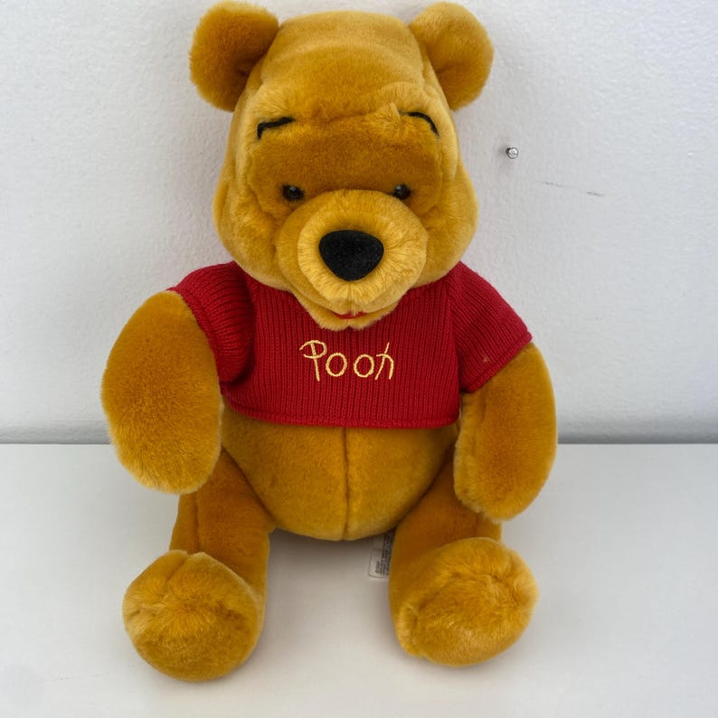 May include: A plush Winnie the Pooh bear, featuring a golden-yellow fur and a red sweater. The sweater has the word "Pooh" embroidered in a gold color. The bear has a black nose and is sitting upright.