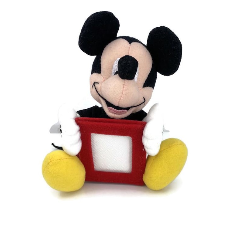 Puede incluir: Un peluche de Mickey Mouse con orejas negras, cara beige y guantes blancos. Mickey sostiene un cuadrado rojo con un centro blanco. El juguete tiene pies amarillos y un cuerpo negro.