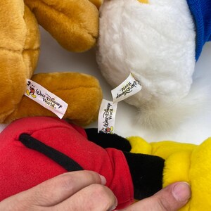 May include: Close-up of several Disney plush toys, including Mickey Mouse, Donald Duck, and a yellow bear. Each toy has a tag that reads "Walt Disney World."