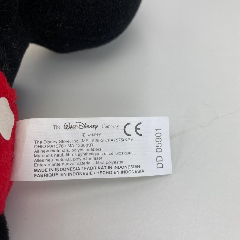 Puede incluir: Una etiqueta blanca con texto negro de The Walt Disney Company, con la marca CE y el n&uacute;mero DD 05901. La etiqueta enumera los materiales y la informaci&oacute;n de fabricaci&oacute;n. La etiqueta est&aacute; adherida a un peluche negro y rojo.