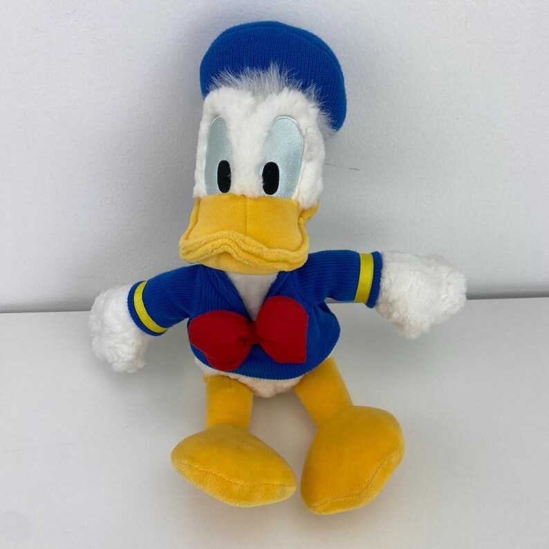 May include: A plush Donald Duck toy. The toy features a blue sailor hat, a blue jacket with yellow accents, a red bow tie, and yellow feet. The duck has a yellow beak, white face, and blue eyes. The toy is made of soft, plush material.