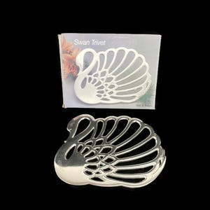 May include: Silver swan-shaped trivet with a cutout design. The trivet is packaged in a white box with the words "Swan Trivet" printed on the front.