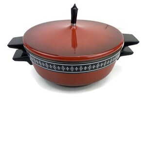 May include: A round, burnt-orange casserole dish with a matching lid. The pot features black side handles and a black lid knob. A black band encircles the upper portion of the pot, decorated with a repeating white geometric diamond pattern.