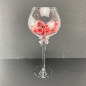 May include: A clear glass vase with a wide bowl and a tall stem. The vase is filled with red, sparkly ornaments.
