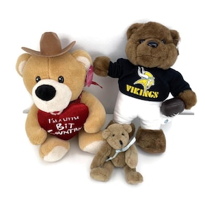 May include: Three plush teddy bears. One tan bear wears a brown cowboy hat and holds a red heart with the text "I'M A LITTLE BIT COUNTRY". Another brown bear wears a black Vikings jersey and white pants, holding a football. A small brown bear is also present.