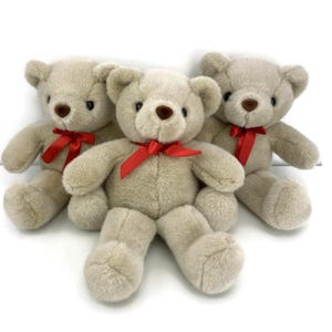 May include: Three plush teddy bears in a light beige color. Each bear has a red ribbon tied in a bow around its neck. The bears have brown eyes and noses, and are sitting upright.