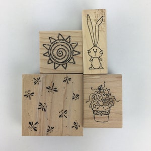 May include: Four wooden stamps with different designs. The first stamp features a sun with a spiral in the center. The second stamp features a rabbit with long ears. The third stamp features a pattern of dragonflies. The fourth stamp features a potted plant with flowers.