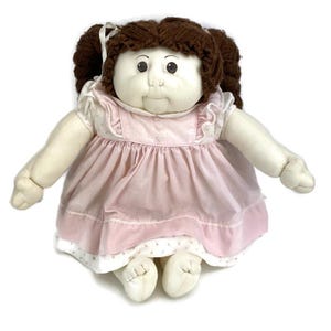 May include: A vintage doll with a soft, plush body and a round face. The doll has brown yarn hair styled in pigtails, and is wearing a pink dress with white trim. The doll's arms and legs are extended, and it has a sweet, innocent expression.