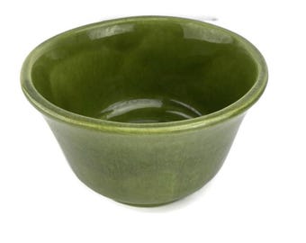 May include: A green ceramic bowl with a glossy finish. The bowl has a rounded shape with a slightly flared rim. The color is a deep, earthy green, and the bowl appears to be handmade.