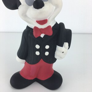 Op de afbeelding: Een zwart-witte keramische figuur van Mickey Mouse die een zwart smoking met een rode strik draagt en een zwarte hoed vasthoudt. De figuur staat op een wit oppervlak.