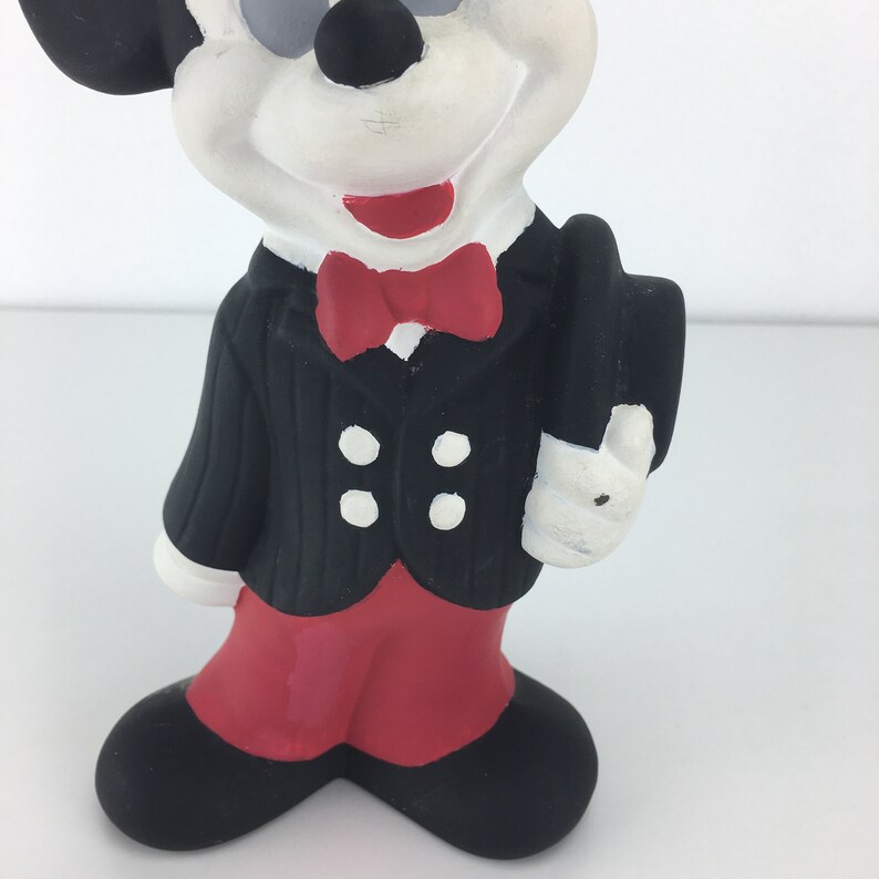Op de afbeelding: Een zwart-witte keramische figuur van Mickey Mouse die een zwart smoking met een rode strik draagt en een zwarte hoed vasthoudt. De figuur staat op een wit oppervlak.