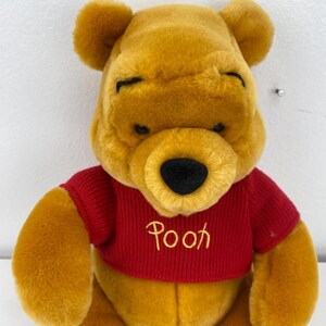 May include: A plush Winnie the Pooh toy in a seated position. The bear is golden yellow with a red sweater. The word "Pooh" is embroidered on the sweater in a cursive font. The bear has closed eyes and a black nose.