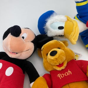 May include: Close-up of plush Disney characters: Mickey Mouse, Donald Duck, and Winnie the Pooh. Mickey Mouse has a black hat and red shorts. Donald Duck has a blue hat and yellow beak. Winnie the Pooh wears a red sweater with "Pooh" embroidered on it.