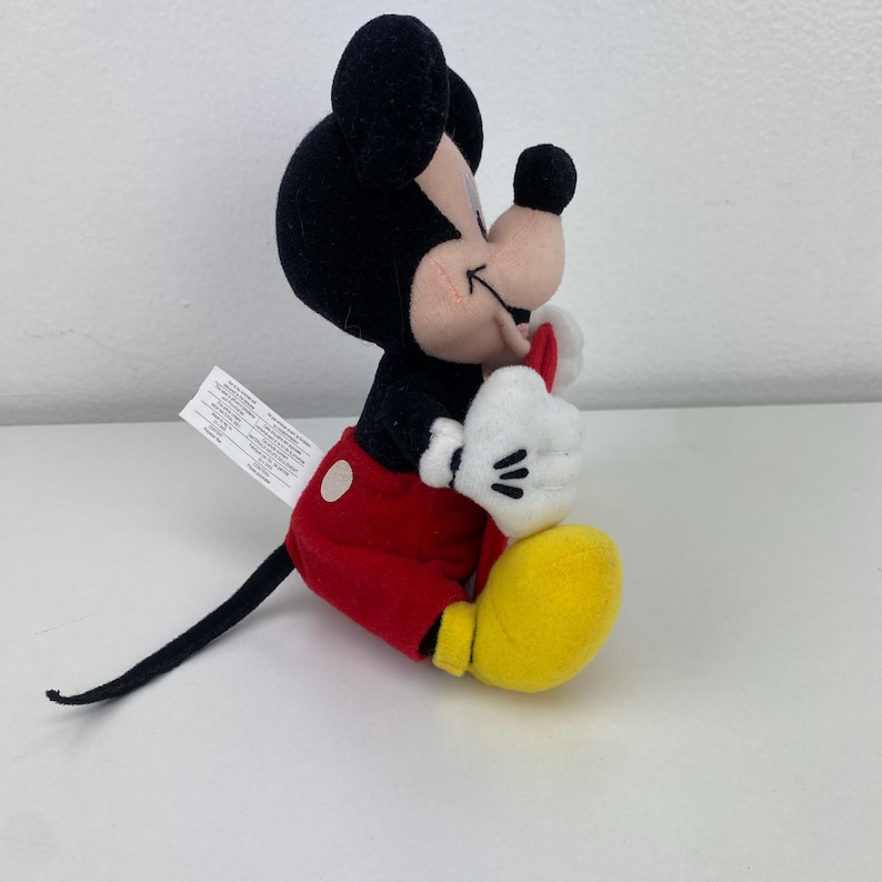 Puede incluir: Un peluche de Mickey Mouse sentado. Mickey lleva pantalones cortos rojos, zapatos amarillos, guantes blancos y una pajarita roja. Sus orejas y cabeza son negras, y su cara es rosa con una nariz negra. Una etiqueta blanca est&aacute; adjunta.