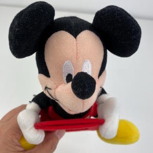 Puede incluir: Un peluche de Mickey Mouse con orejas negras, una nariz negra y un atuendo rojo. El juguete tiene guantes blancos y zapatos amarillos. El juguete sostiene un objeto rojo.