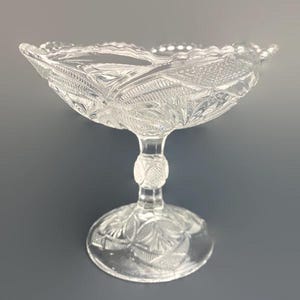 Könnte beinhalten: Klarer Glasfußschale mit aufwendigen geprägten floralen und geometrischen Mustern. Die Schale hat einen gewellten Rand und einen runden Sockel. Ein dekoratives Stück zum Servieren oder Ausstellen.