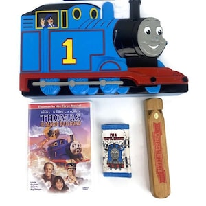 Puede incluir: Un tren de juguete azul y negro con un silbato, un DVD de Thomas y el Ferrocarril Mágico, una bolsa de tela azul con el texto "Soy una locomotora útil", y un silbato de madera con el texto "El silbato Dixie".