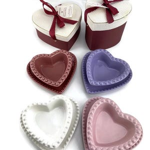 Op de afbeelding: Een assortiment hartvormige items. Twee hartvormige geschenkdozen met crèmekleurige deksels en bordeauxrode bodems zijn vastgebonden met bordeauxrode linten. Hieronder bevinden zich vier sets hartvormige schalen in bordeauxrood, paars, wit en roze, elk met een parelrand.
