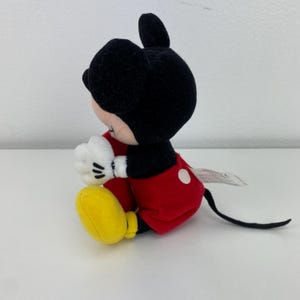 Puede incluir: Un peluche de Mickey Mouse en posici&oacute;n sentada. El juguete presenta una cabeza y orejas negras, un cuerpo rojo, guantes blancos y zapatos amarillos. El juguete tiene una cola negra y una etiqueta blanca.