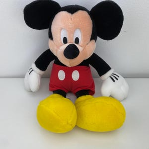 May include: A plush Mickey Mouse doll with a black head and ears, a tan face, and a black nose. It wears red shorts with white buttons, white gloves, and yellow shoes. The doll is approximately 18 inches tall.