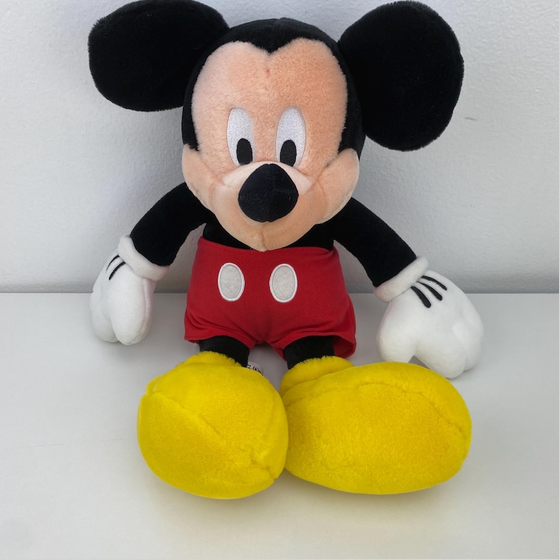 May include: A plush Mickey Mouse doll with a black head and ears, a tan face, and a black nose. It wears red shorts with white buttons, white gloves, and yellow shoes. The doll is approximately 18 inches tall.