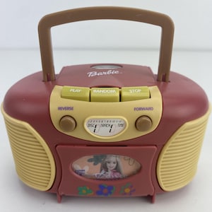 May include: A maroon and beige portable Barbie boombox with a top handle. The device features buttons labelled Play, Random, Stop, Reverse, and Forward, along with a digital display showing Disc, Track, and Time. A central window displays a Barbie graphic.