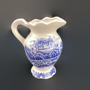 May include: White ceramic pitcher with a blue and white landscape design. The pitcher features a handle and a scalloped rim. The blue design depicts a castle and surrounding scenery. The base of the pitcher has a floral pattern.