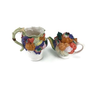 May include: A white ceramic teapot and creamer set decorated with a colourful assortment of fruits and vegetables, including pumpkins, grapes, and corn. The set is perfect for a autumn-themed tea party.