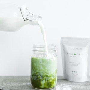 Pu&ograve; includere: Un barattolo di vetro pieno di una bevanda al matcha verde con latte che viene versato al suo interno da una bottiglia di vetro. Il barattolo &egrave; appoggiato su una superficie di legno con cubetti di ghiaccio e un cucchiaio con polvere di matcha.