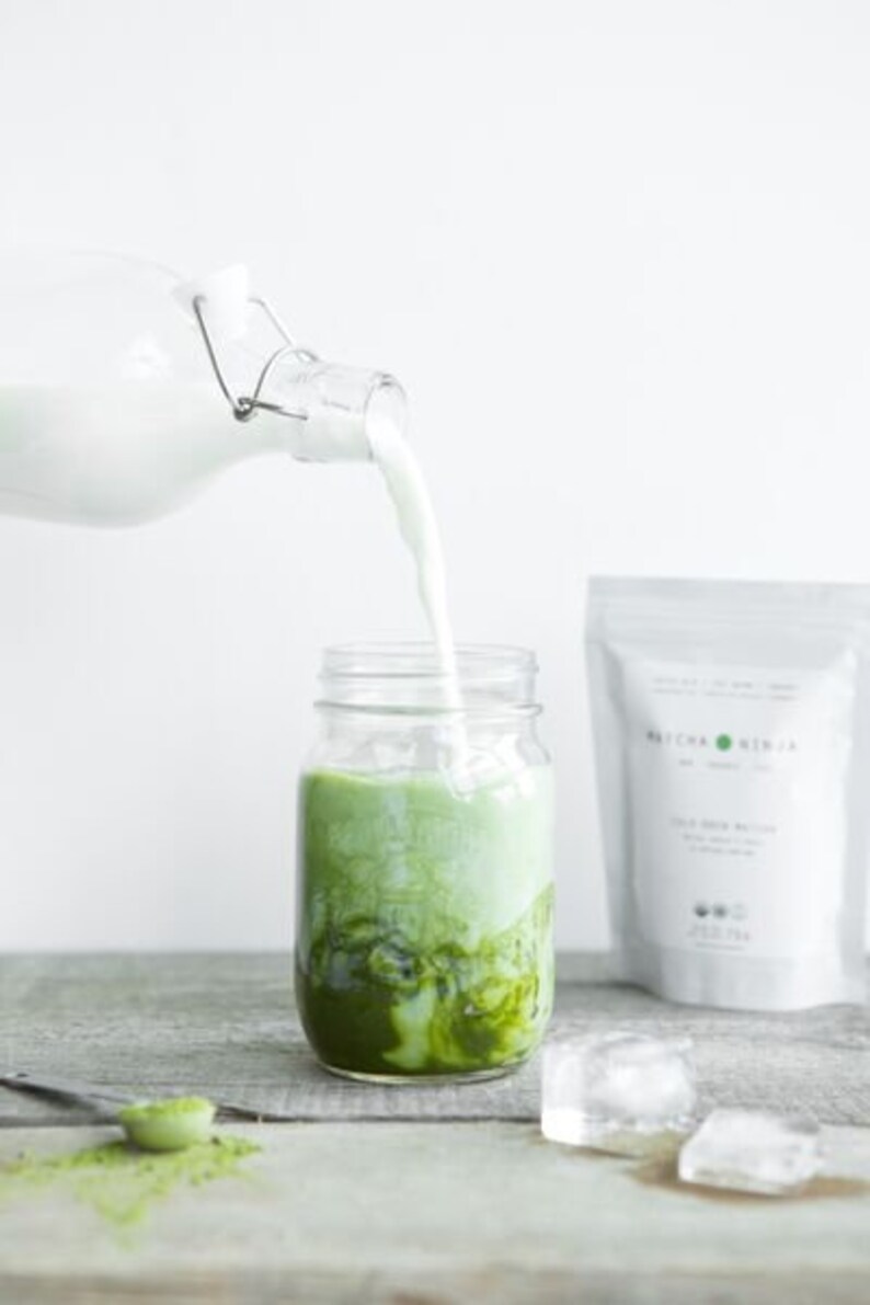 Pu&ograve; includere: Un barattolo di vetro pieno di una bevanda al matcha verde con latte che viene versato al suo interno da una bottiglia di vetro. Il barattolo &egrave; appoggiato su una superficie di legno con cubetti di ghiaccio e un cucchiaio con polvere di matcha.