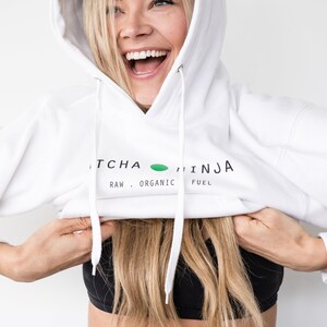 Pu&ograve; includere: Una felpa con cappuccio bianca con la scritta "MATCHA NINJA RAW. ORGANIC FUEL" stampata sul davanti. La modella indossa la felpa con cappuccio e la tira gi&ugrave; per rivelare un reggiseno sportivo nero.