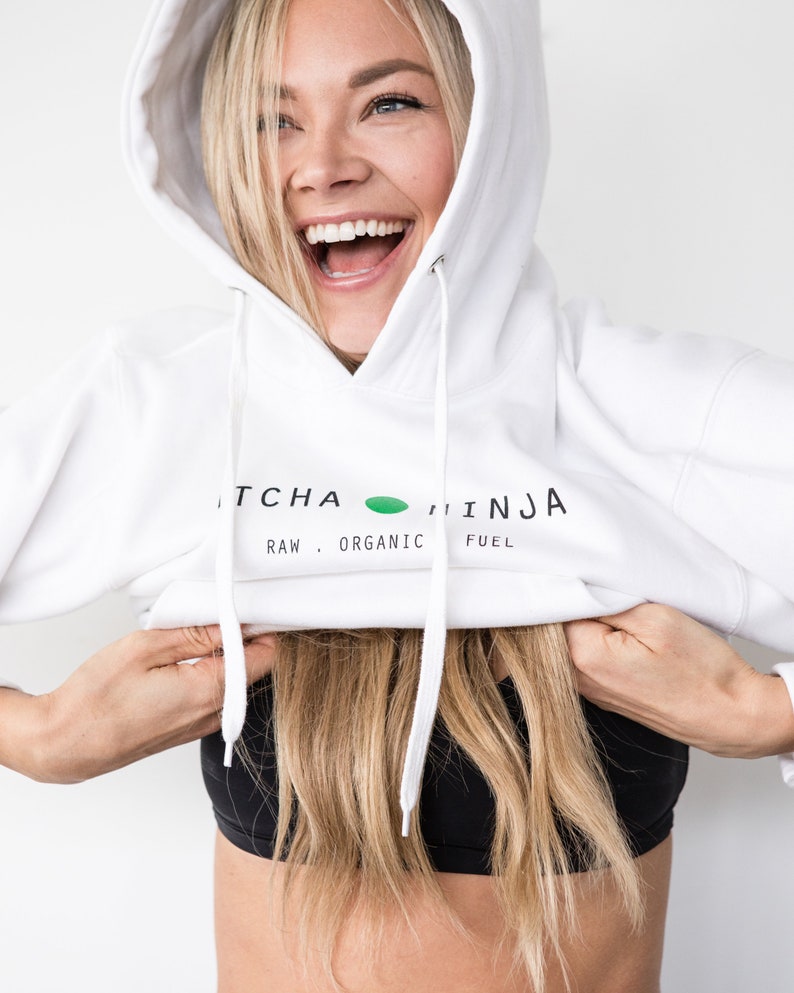 Pu&ograve; includere: Una felpa con cappuccio bianca con la scritta "MATCHA NINJA RAW. ORGANIC FUEL" stampata sul davanti. La modella indossa la felpa con cappuccio e la tira gi&ugrave; per rivelare un reggiseno sportivo nero.