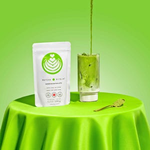 Pu&ograve; includere: Un latte matcha verde viene versato in un bicchiere con un bordo bianco. Il bicchiere &egrave; su una tovaglia verde. Una bustina bianca con il testo "MATCHA NINJA SUPERFOOD MATCHA LATTE" &egrave; sul tavolo.