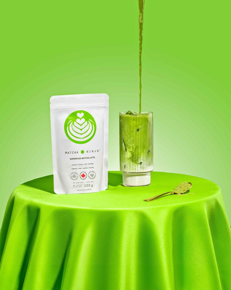 Pu&ograve; includere: Un latte matcha verde viene versato in un bicchiere con un bordo bianco. Il bicchiere &egrave; su una tovaglia verde. Una bustina bianca con il testo "MATCHA NINJA SUPERFOOD MATCHA LATTE" &egrave; sul tavolo.