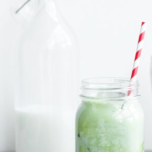 Pu&ograve; includere: Un barattolo di vetro con una cannuccia a strisce rosse e bianche, riempito di un latte matcha verde. Il barattolo &egrave; appoggiato su una superficie di legno accanto a una bottiglia di vetro trasparente piena di latte.