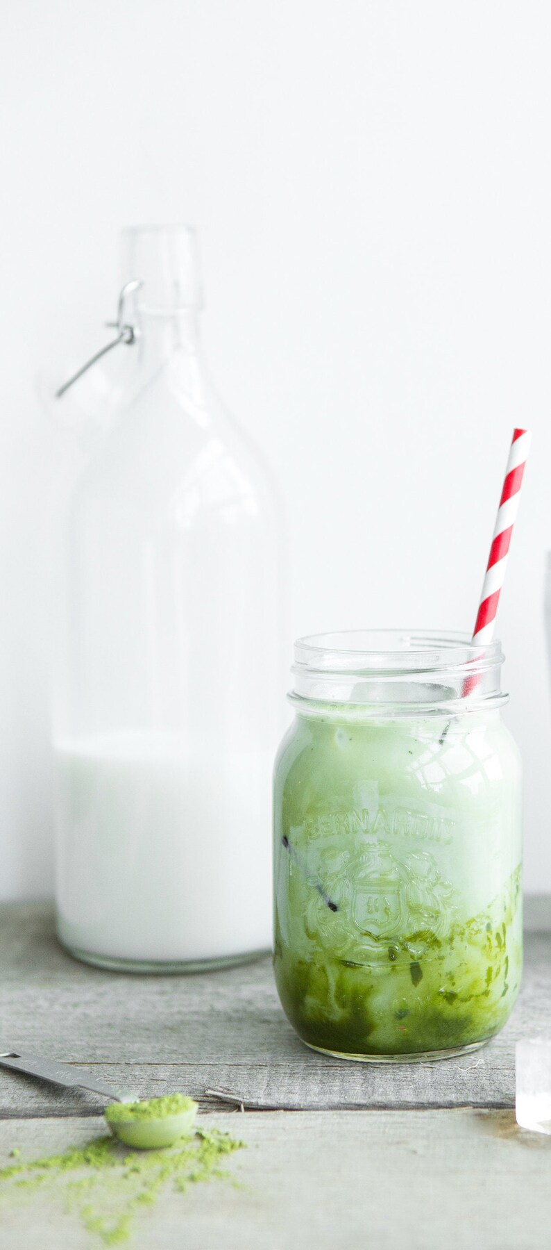 Pu&ograve; includere: Un barattolo di vetro con una cannuccia a strisce rosse e bianche, riempito di un latte matcha verde. Il barattolo &egrave; appoggiato su una superficie di legno accanto a una bottiglia di vetro trasparente piena di latte.