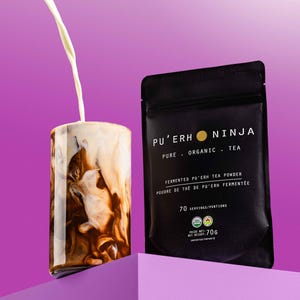 May include: A black pouch with white text that reads "PU'ERH NINJA PURE ORGANIC TEA FERMENTED PU'ERH TEA POWDER POUDRE DE THE DE PU'ERH FERMENTEE 70 SERVINGS/PORTIONS POIDS NET/NET WEIGHT 70G" and a yellow circle with a black outline. The pouch is sitting on a purple surface. A glass with a brown and white liquid is in the background.