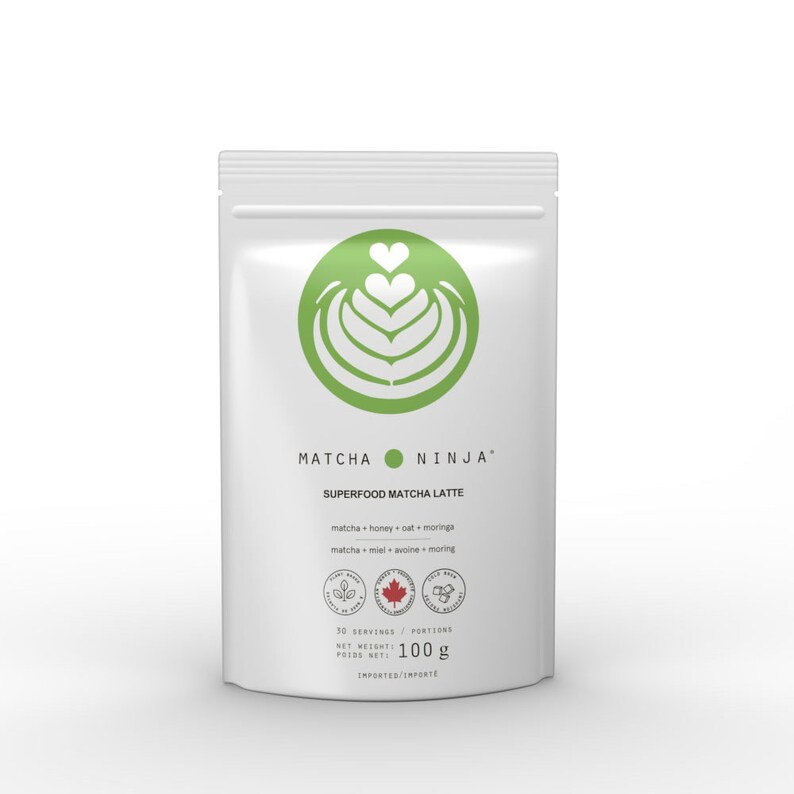 Pu&ograve; includere: Una busta bianca con un logo verde a forma di cuore con un design a spirale. Il testo "MATCHA NINJA SUPERFOOD MATCHA LATTE" &egrave; stampato sulla busta. Gli ingredienti sono elencati come "matcha + honey + oat + moringa" e "matcha + miel + avoine + moringa". La busta contiene 100 grammi di polvere di matcha.