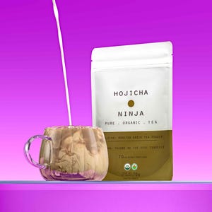 Può includere: Una busta di polvere di tè verde tostato hojicha biologico di Ninja. La busta è marrone con testo bianco e un piccolo cerchio al centro. Il testo recita "Hojicha Ninja Pure Organic Tea" e "Uichiai Roasted Green Tea Powder". La busta è su una superficie viola con un bicchiere di latte che viene versato al suo interno.