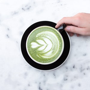 Pu&ograve; includere: Una tazza di latte matcha verde con un disegno di schiuma bianca in cima, appoggiata su un piattino nero. La tazza &egrave; tenuta da una mano.