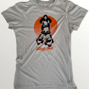 May include: A light gray t-shirt with a black and white illustration of a woman roller skating in front of an orange circle. The text "Skate Ink" is printed below the illustration.