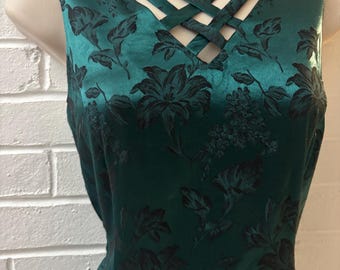May include: A sleeveless dress in a deep emerald green satin fabric featuring a black floral pattern. The neckline is accented with a crisscross strap design, creating a geometric cutout effect across the chest area of the garment.